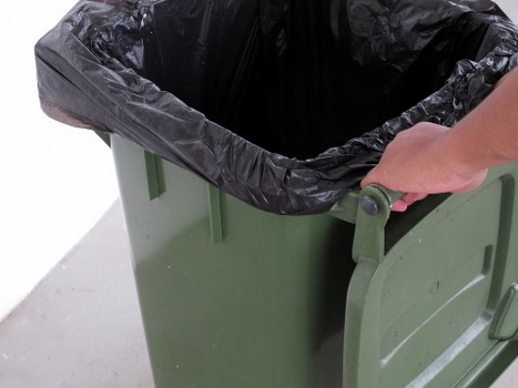 A close-up of a green plastic wheeled rubbish bin with a black plastic liner draped over the top. A person's hand is seen gripping the handle of the bin, which is positioned outdoors on a paved surface. The background is plain and uncluttered, focusing on the bin's simple design and the action of holding it, typical of waste collection or rubbish removal activities in a residential or commercial setting. The scene illustrates standard rubbish disposal equipment used by waste management services such as House Clearance Hemel Hempstead to facilitate rubbish removal in the local area.