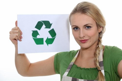 A woman with blonde hair styled in a side braid, wearing a green top and grey apron, is holding a white sign with a large green recycling symbol composed of three arrows forming a triangle. She is standing outdoors, possibly on a driveway or pavement, with a neutral background. The image emphasizes environmentally conscious waste management and recycling practices relevant to rubbish removal services in Hemel Hempstead, as indicated by the context of house clearance and waste disposal. The woman appears to be presenting the recycling message clearly, supporting local efforts in sustainable rubbish disposal managed by House Clearance Hemel Hempstead. The setting is well-lit with natural light, and the focus is on the woman and the recycling symbol, highlighting the importance of responsible waste separation and recycling in the local area.