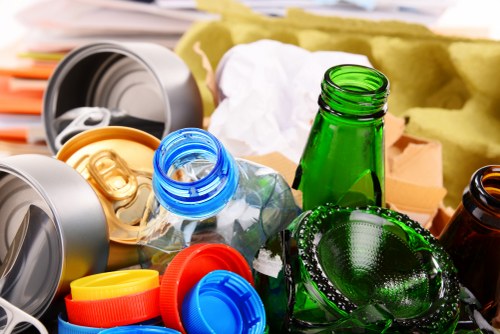 A cluttered assortment of recyclable bottles and containers, including clear plastic, green glass, and amber glass, with some caps removed or loose, is piled together on a surface likely outdoors, in a setting that appears to be part of waste collection or disposal. The bottles vary in shape and size, with textured surfaces visible on the green glass bottle, and some are partially obscured by other items, such as lids and wrappers. In the background, there is a yellowish material, possibly packaging or insulation, along with a blurred object resembling a storage container or trash bin, indicating an environment where rubbish or waste materials are being sorted or prepared for collection. The scene reflects the type of waste management services provided by House Clearance Hemel Hempstead, serving local households in the Hemel Hempstead area, with a focus on the collection and disposal of recyclable waste materials in a professional and organized manner.