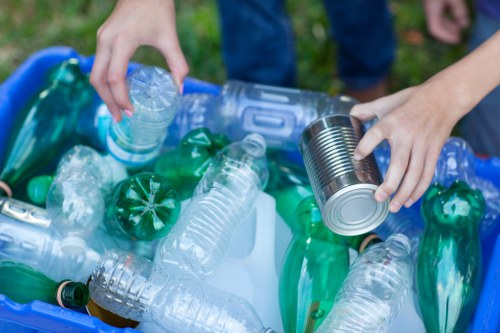 A close-up of a person's hands sorting recyclable plastic bottles and cans into a collection bin, with the background showing a grassy outdoor area. The bottles are of various sizes and transparent or green in color, some with caps still on, and are made of clear or green plastic. One hand is holding a plastic bottle, while the other is holding a tin can, both positioned above the gathered collection. The scene reflects an environment of responsible waste segregation, typical of rubbish removal and recycling efforts, possibly in a residential or community setting near Hemel Hempstead. The collection area is outdoors, with natural daylight illuminating the scene, emphasizing the textures of the plastic and metal objects. This accurate depiction supports the importance of proper rubbish disposal and recycling services offered by companies like House Clearance Hemel Hempstead.