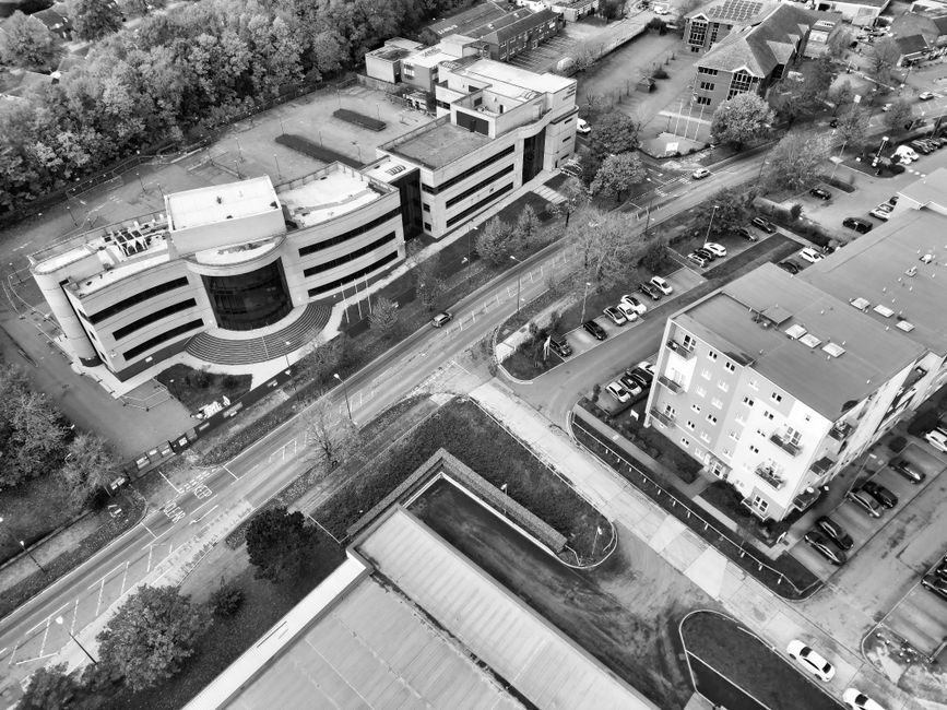 An aerial black-and-white photograph shows a densely built urban area with a mixture of commercial and residential buildings. In the foreground, there is a large, flat-roofed retail or warehouse compl