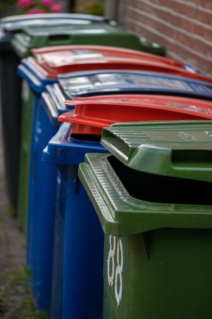 A row of four dark brown wooden bins, each with a sloped flat roof, are positioned side by side on a grassy area with some scattered fallen leaves. In front of the bins, there are various discarded it